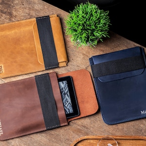 Può includere: Tre custodie in pelle per e-reader in marrone, beige e blu navy, ciascuna con chiusura a fascia elastica nera. La custodia marrone è aperta, rivelando un e-reader. Le custodie beige e blu navy hanno delle iniziali. Una piccola pianta verde è nelle vicinanze.