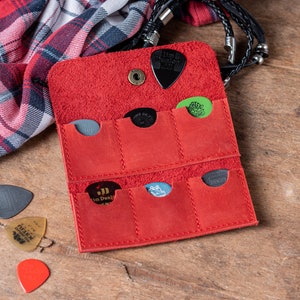 May include: Red leather guitar pick holder with six slots for storing picks. The holder is open and several picks are visible, including a green pick with a skull design, a black pick with the text "Jim Dunlop", and a black pick with the text "The Grip 1.0".