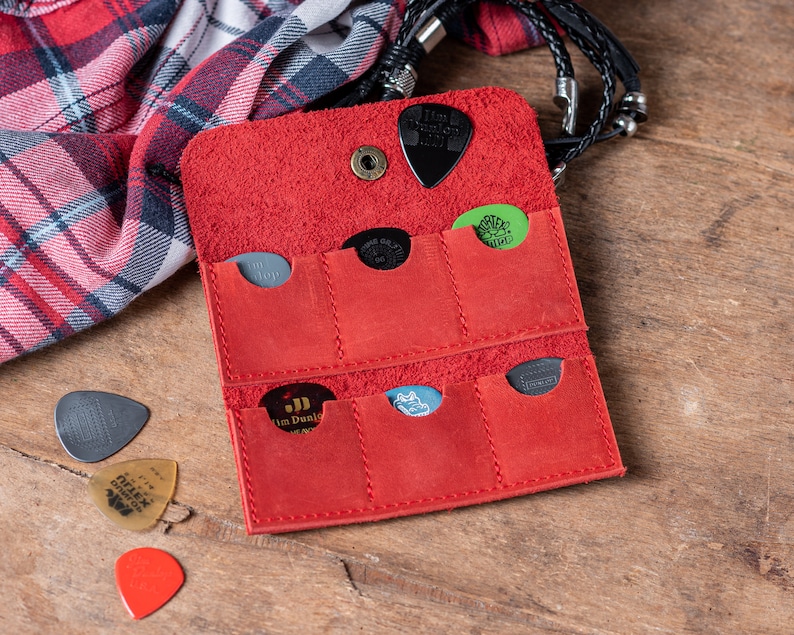 May include: Red leather guitar pick holder with six slots for storing picks. The holder is open and several picks are visible, including a green pick with a skull design, a black pick with the text "Jim Dunlop", and a black pick with the text "The Grip 1.0".