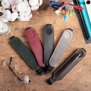 K&ouml;nnte beinhalten: F&uuml;nf Leder-Stift-Etuis in verschiedenen Farben, darunter Gr&uuml;n, Rot, Grau und Schwarz, sowie ein braunes Leder-Stift-Etui mit einem silbernen Stift. Die Stiftetuis haben einen Trageriemen.