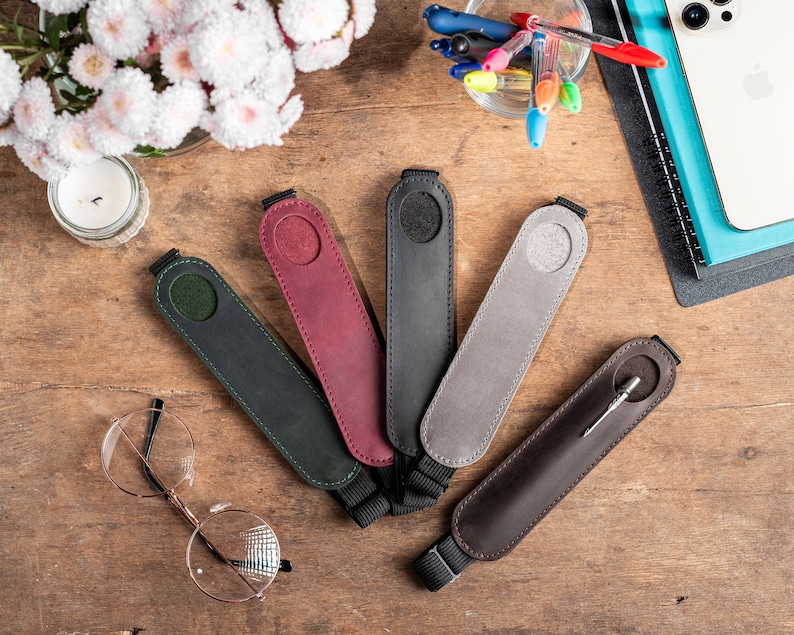 K&ouml;nnte beinhalten: F&uuml;nf Leder-Stift-Etuis in verschiedenen Farben, darunter Gr&uuml;n, Rot, Grau und Schwarz, sowie ein braunes Leder-Stift-Etui mit einem silbernen Stift. Die Stiftetuis haben einen Trageriemen.