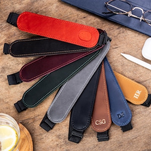 May include: A collection of leather pen holders in various colors, including red, black, and blue. Each holder has a stitched design and a black elastic strap. Some holders feature initials. The items are arranged on a wooden surface.