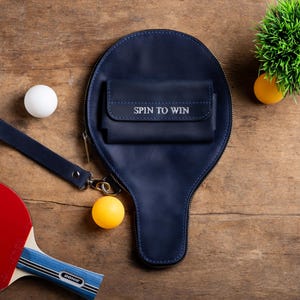 May include: A navy blue ping pong paddle case with a small wallet that reads "SPIN TO WIN". A red and blue ping pong paddle, two ping pong balls, and a small green plant are also visible.
