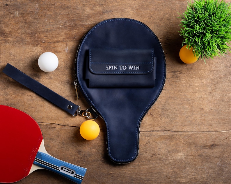 May include: A navy blue ping pong paddle case with a small wallet that reads "SPIN TO WIN". A red and blue ping pong paddle, two ping pong balls, and a small green plant are also visible.