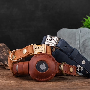 May include: Two leather dog collars with engraved name tags and phone numbers. The collars are brown and navy blue. The collars are on a wooden surface with a green plant in the background.