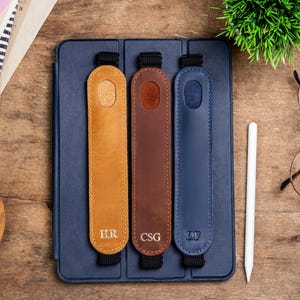 May include: Three leather tablet holders in tan, brown, and navy blue, each with a black strap and initials. The holders are on a navy blue tablet case, with a white stylus and glasses on a wooden surface.