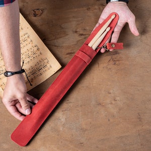 Puede incluir: Un porta baquetas de cuero rojo con cierre de cremallera y un peque&ntilde;o lazo de cuero. Dos baquetas de madera est&aacute;n dentro del porta baquetas.