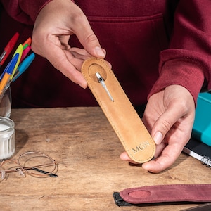 K&ouml;nnte beinhalten: Ein brauner Leder-Stift-Halter mit einem silbernen Stift im Inneren. Der Halter hat die Initialen "MCN" unten eingepr&auml;gt.
