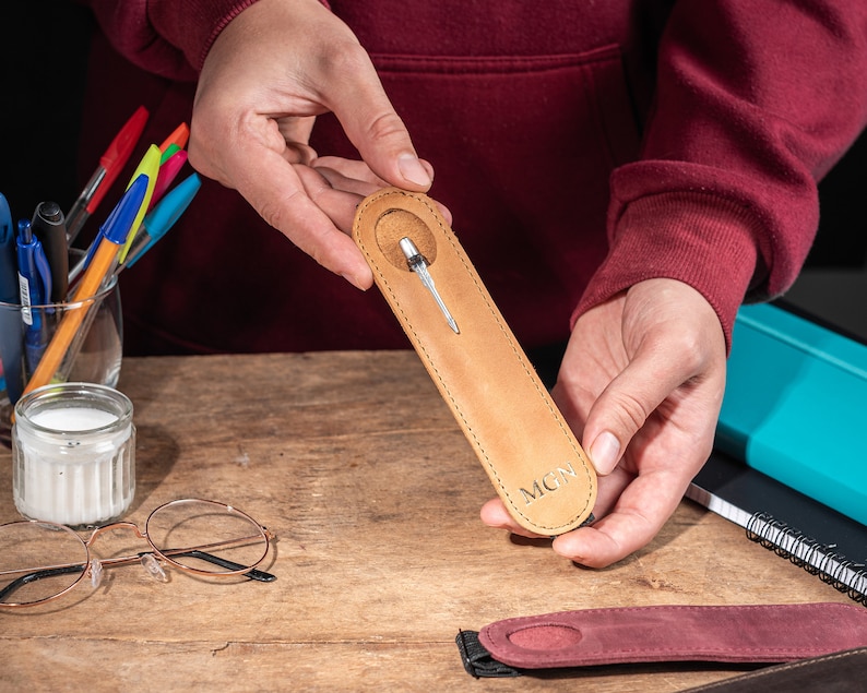 K&ouml;nnte beinhalten: Ein brauner Leder-Stift-Halter mit einem silbernen Stift im Inneren. Der Halter hat die Initialen "MCN" unten eingepr&auml;gt.