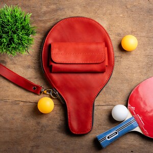 May include: A red leather ping pong paddle-shaped bag with a small wallet attached. The bag has a wrist strap and a zipper. Two yellow and one white ping pong balls are next to the bag, along with a red and blue ping pong paddle and a small green plant.