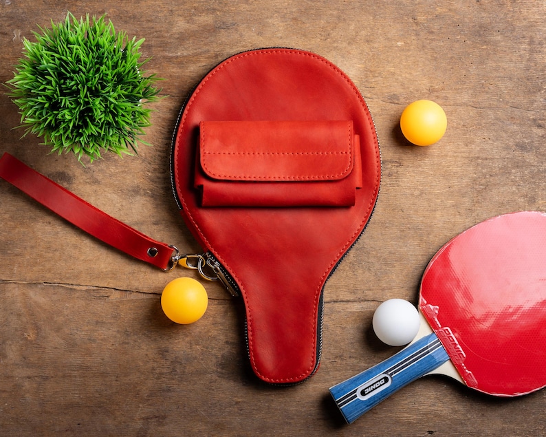 May include: A red leather ping pong paddle-shaped bag with a small wallet attached. The bag has a wrist strap and a zipper. Two yellow and one white ping pong balls are next to the bag, along with a red and blue ping pong paddle and a small green plant.