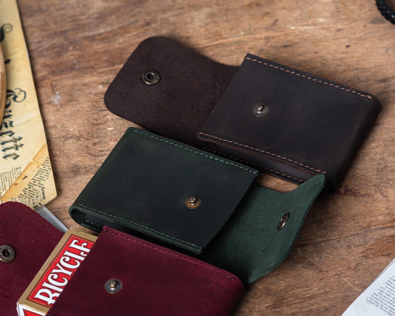 May include: Three leather card cases in different colors: brown, green, and burgundy. Each case has a snap closure and a deck of cards is visible in the burgundy case.