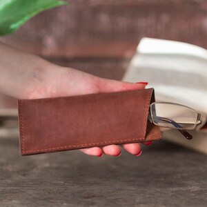 May include: A brown leather glasses case with a pair of glasses inside. The case is closed and held in a hand.
