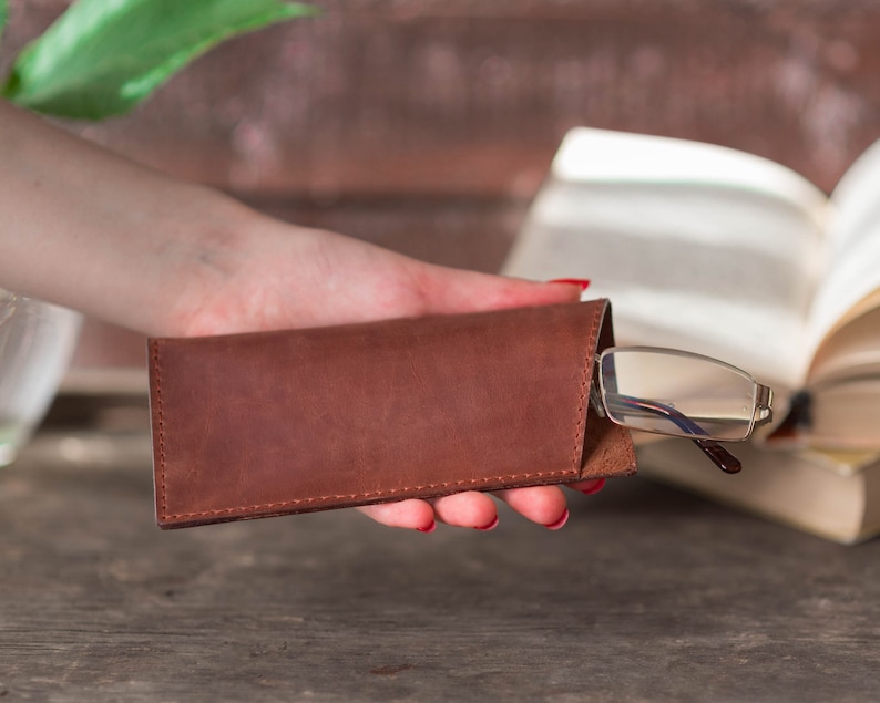 May include: A brown leather glasses case with a pair of glasses inside. The case is closed and held in a hand.