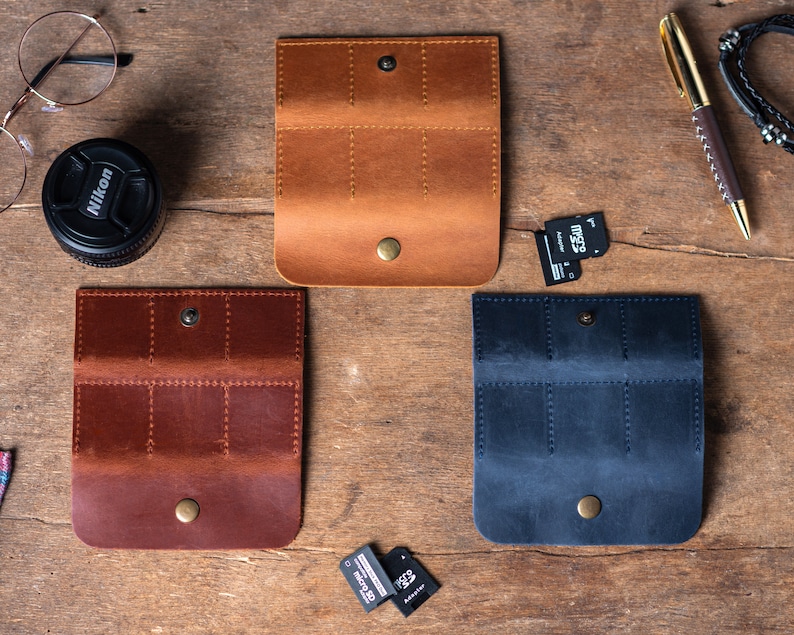 May include: Three leather cases for memory cards. The cases are brown, dark brown, and blue. Each case has multiple slots for memory cards. The cases are on a wooden surface.
