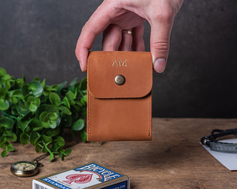 May include: A brown leather card holder with a flap and a button closure. The holder has the initials "AM" embossed in gold on the front.