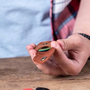 以下が含まれることがあります： カラフルなギターピックが収納された、茶色の革製のピックホルダー。フラップを開けるとピックが見えます。