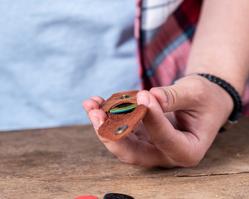 以下が含まれることがあります： カラフルなギターピックが収納された、茶色の革製のピックホルダー。フラップを開けるとピックが見えます。