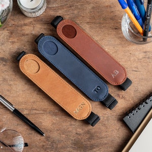 K&ouml;nnte beinhalten: Drei Leder-Stift-Halter mit personalisierten Initialen. Die Halter sind braun, blau und beige mit schwarzen Gummib&auml;ndern. Die Initialen sind MGN, TO und AB.