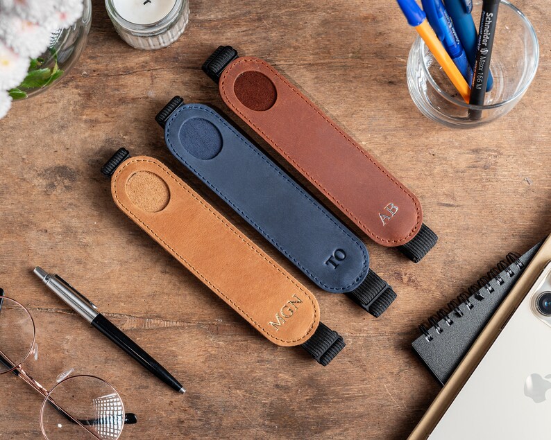 K&ouml;nnte beinhalten: Drei Leder-Stift-Halter mit personalisierten Initialen. Die Halter sind braun, blau und beige mit schwarzen Gummib&auml;ndern. Die Initialen sind MGN, TO und AB.