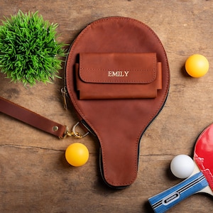 May include: A brown leather ping pong paddle-shaped bag with a matching wallet, personalized with the name "EMILY". The bag has a wrist strap and a gold zipper. Two yellow and one white ping pong balls are next to a red and blue ping pong paddle.