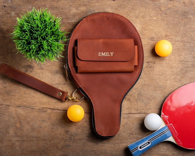 May include: A brown leather ping pong paddle-shaped bag with a matching wallet, personalized with the name "EMILY". The bag has a wrist strap and a gold zipper. Two yellow and one white ping pong balls are next to a red and blue ping pong paddle.