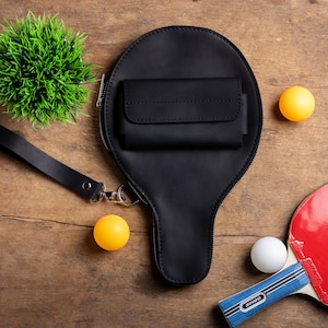May include: A black leather ping pong paddle-shaped bag with a small wallet attached, a wrist strap, and a zipper. Two yellow and one white ping pong balls are scattered around the bag, along with a red and blue ping pong paddle and a small green plant.
