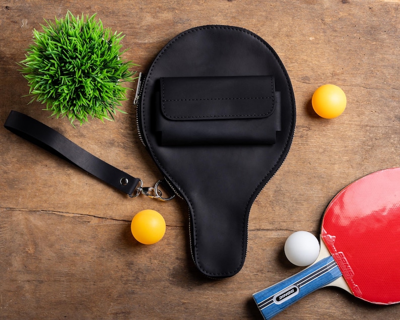 May include: A black leather ping pong paddle-shaped bag with a small wallet attached, a wrist strap, and a zipper. Two yellow and one white ping pong balls are scattered around the bag, along with a red and blue ping pong paddle and a small green plant.