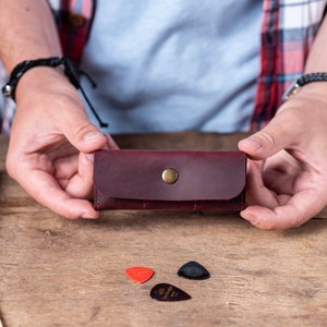 May include: A burgundy leather guitar pick holder with a brass button closure. The holder is being held by a person's hands. There are three guitar picks on the table, two red and one black.