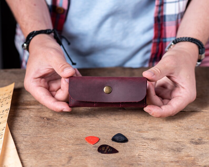 May include: A burgundy leather guitar pick holder with a brass button closure. The holder is being held by a person's hands. There are three guitar picks on the table, two red and one black.