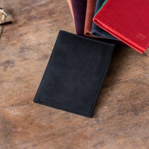 May include: A black leather wallet with a stitched edge, next to a stack of other wallets in various colors, including red, brown, and green. The red wallet has the initials "SF" embossed on the front.