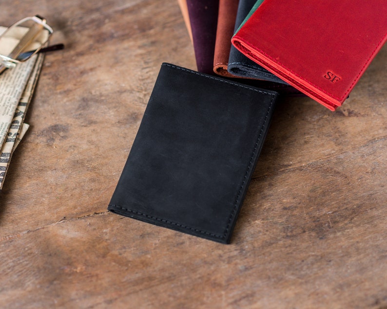 May include: A black leather wallet with a stitched edge, next to a stack of other wallets in various colors, including red, brown, and green. The red wallet has the initials "SF" embossed on the front.