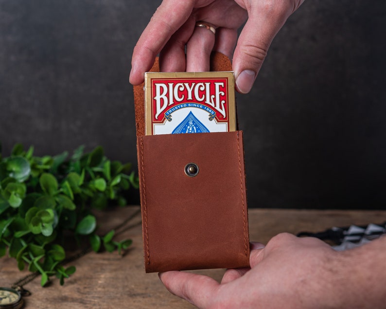 May include: A brown leather card holder with a snap closure. A deck of Bicycle playing cards is partially inserted into the holder.