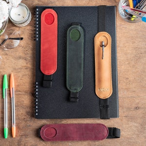 K&ouml;nnte beinhalten: Vier Leder-Stift-Halter mit Gummib&auml;ndern. Die Halter sind in Rot, Gr&uuml;n, Braun und Burgunderrot. Der braune Halter hat die Buchstaben "MGN" gepr&auml;gt.