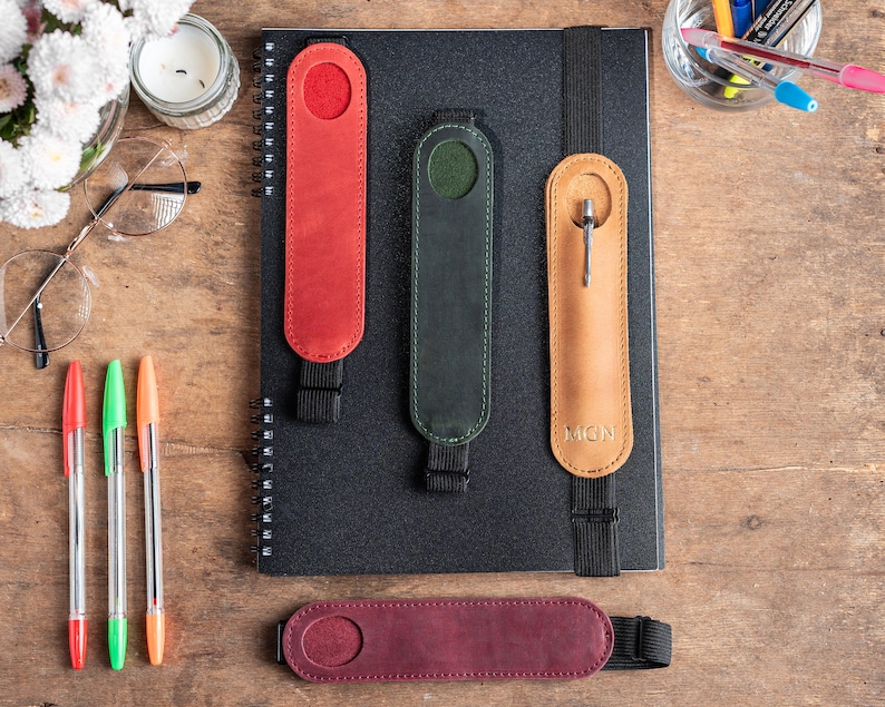 K&ouml;nnte beinhalten: Vier Leder-Stift-Halter mit Gummib&auml;ndern. Die Halter sind in Rot, Gr&uuml;n, Braun und Burgunderrot. Der braune Halter hat die Buchstaben "MGN" gepr&auml;gt.