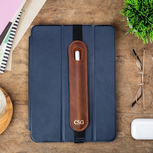 May include: A navy blue tablet case with a brown leather stylus holder, featuring the initials "CSG". The case is secured with a black elastic band. Additional items include a pair of gold-rimmed glasses, a white wireless earbud case, and a small green plant.
