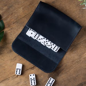 May include: A dark navy blue leather roll-up pouch containing a set of white dominoes with black pips. Three additional white dominoes with black pips are positioned outside the pouch, displaying various numerical combinations on their surfaces.