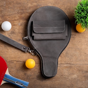May include: A gray leather ping pong paddle case with a wrist strap and a small pocket. The case is zipped closed and has white stitching. A ping pong paddle, two ping pong balls, and a small green plant are also visible.
