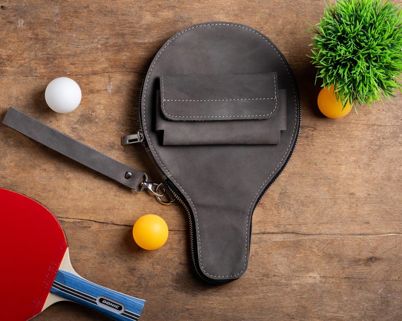 May include: A gray leather ping pong paddle case with a wrist strap and a small pocket. The case is zipped closed and has white stitching. A ping pong paddle, two ping pong balls, and a small green plant are also visible.