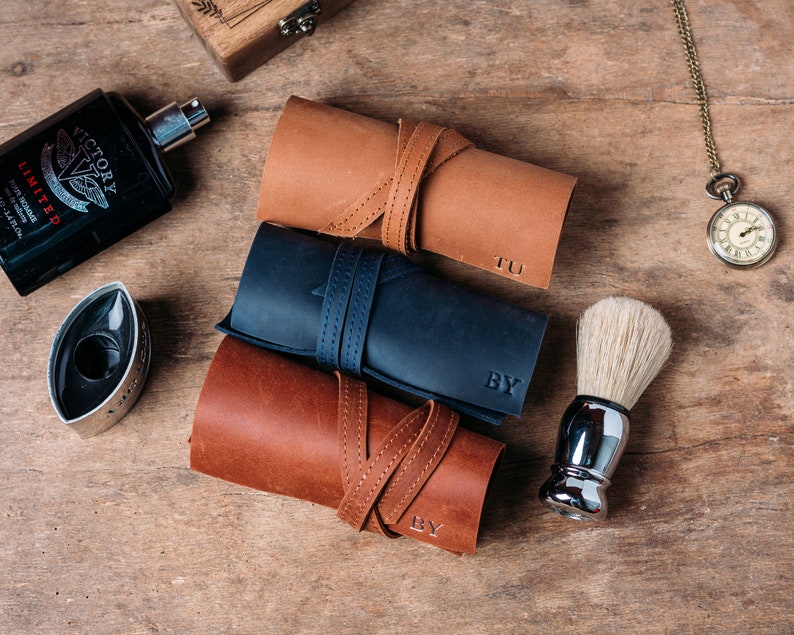 May include: Three rolled leather cases with personalized initials, "TU", "BY", and "BY",  in dark brown, navy blue, and light brown. A silver shaving brush and a bottle of cologne are also pictured.