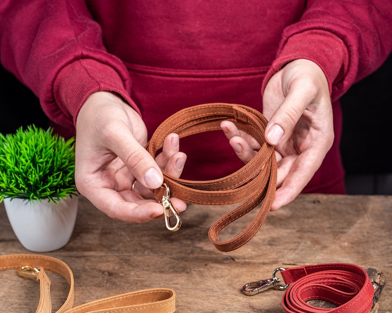 May include: A brown leather strap with a gold metal clasp. The strap is folded in a circle and held in someone's hands.