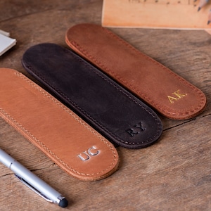 May include: Three leather pen holders with different initials, "UC", "RY", and "AK", are arranged on a wooden surface. The pen holders are brown, black, and tan.