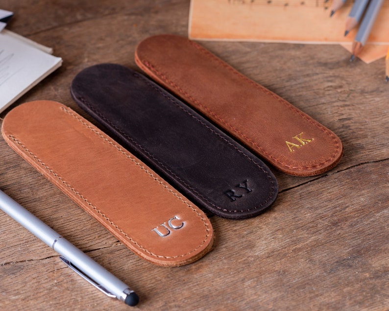 May include: Three leather pen holders with different initials, "UC", "RY", and "AK", are arranged on a wooden surface. The pen holders are brown, black, and tan.