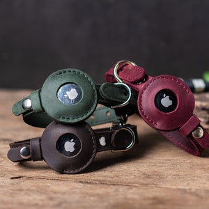 Op de afbeelding: Drie leren AirTag-houders in groen, bruin en bordeauxrood met een Apple-logo op elk. De houders zijn ontworpen om aan een halsband of sleutelhanger te worden bevestigd.