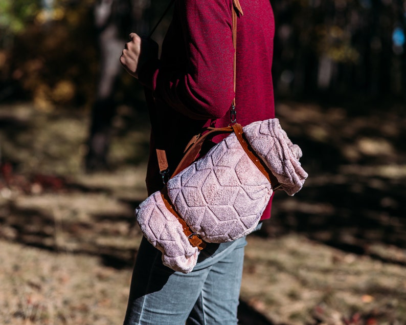 May include: A person wearing a red shirt carries a pink and brown patterned blanket strapped to their shoulder with a brown leather strap.
