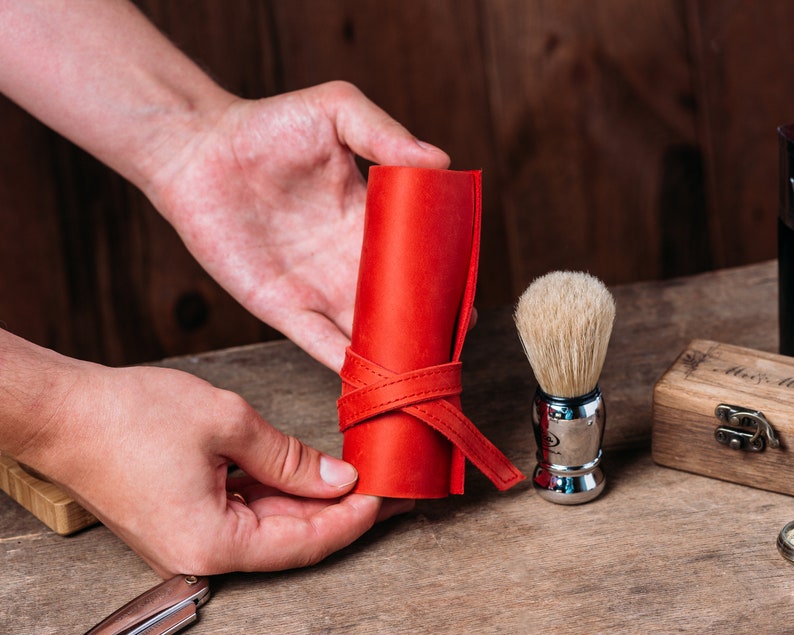 May include: A red leather shaving brush roll with a leather strap closure. The roll is being held by a person's hands. A silver shaving brush with white bristles is in the background.