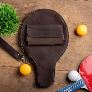 May include: A brown leather paddle-shaped bag with a small pocket, a wrist strap, and a zipper. Two yellow ping pong balls, a white ping pong ball, and a red and blue ping pong paddle are also visible on a wooden surface.