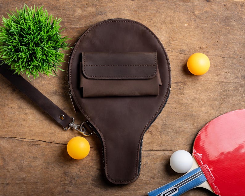 May include: A brown leather paddle-shaped bag with a small pocket, a wrist strap, and a zipper. Two yellow ping pong balls, a white ping pong ball, and a red and blue ping pong paddle are also visible on a wooden surface.