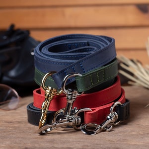 May include: Three leather dog leashes in navy blue, green, and red, stacked on top of each other. Each leash has a silver clip with a gold accent.