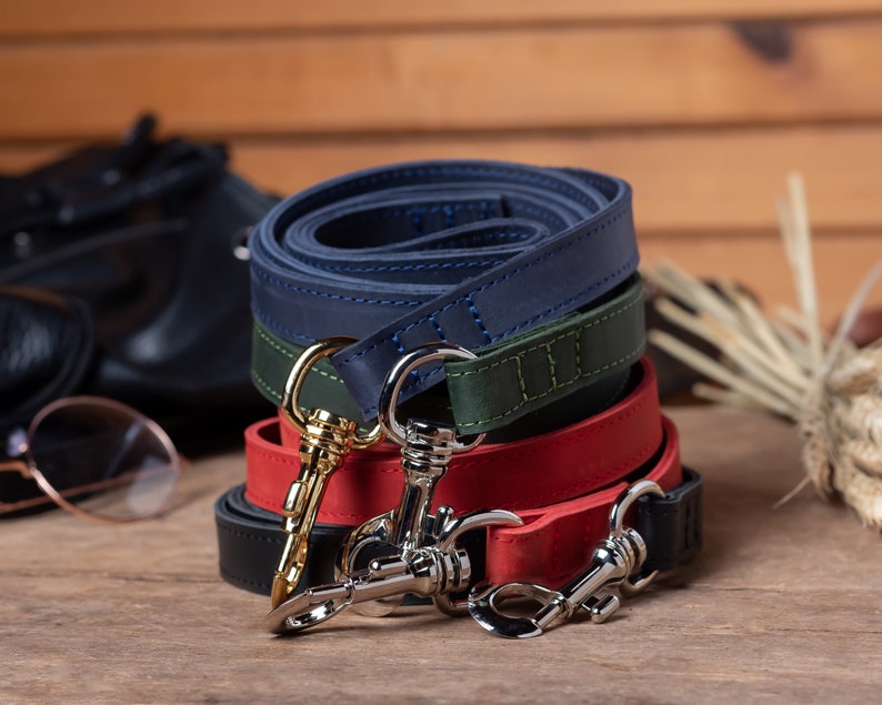 May include: Three leather dog leashes in navy blue, green, and red, stacked on top of each other. Each leash has a silver clip with a gold accent.
